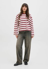 Een vrouw staat tegen een effen achtergrond, gekleed in een gestreepte rood-witte longsleeve, losse grijze broek en zwarte schoenen.