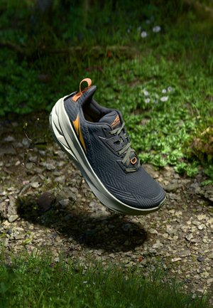 Chaussure de sport noire avec une tige texturée, des accents orange et une semelle blanche. Caractéristiques d'une tige avec une boucle pour un enfilage facile. Flottant au-dessus d'un sol en gravier.