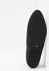 Semelle plate noire de chaussure avec la pointure 37, bout pointu et dessus en tissu texturé, posée devant un fond blanc d’angle.
