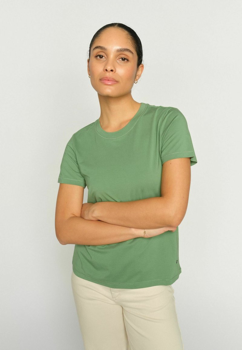 Vrouw met gekruiste armen, gekleed in een groen T-shirt met korte mouwen en beige broek, staand tegen een eenvoudige lichte achtergrond.
