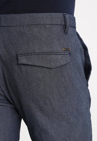 Navyblå byxor i en texturerad tyg med små prickiga mönster, en bakficka och ett guldaccent på fickans söm.