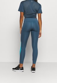 Kvinna står med ryggen mot oss, iklädd blå leggings med en turkos rand och svarta och vita Nike-sneakers.