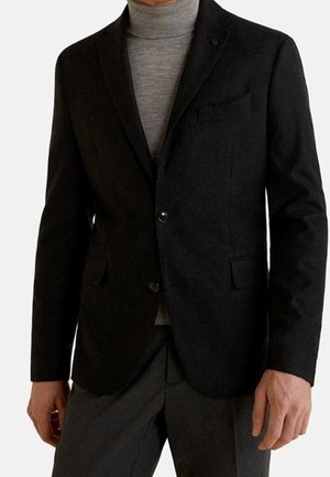 Blazer negro a medida usado sobre un jersey de cuello alto gris claro con pantalones gris oscuro, visto desde los hombros hasta la mitad del muslo.