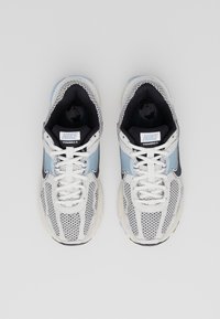 Par de zapatillas de running Nike Vomero 5 en blanco y azul con swooshes negros, cordones blancos y detalles de malla, vistas desde arriba sobre un fondo gris.