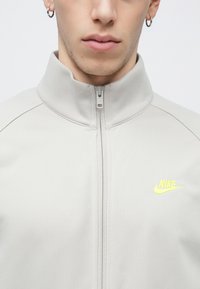 Ανοιχτό γκρι μπουφάν με φερμουάρ και ψηλό γιακά, με κίτρινο λογότυπο Nike. Λείο υλικό και άψογη εφαρμογή με αντιθετικές ραφές.