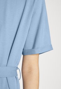 Blusa azul clara de manga curta com punhos dobrados e laço na cintura, feita de um tecido suave, com leve drapeado e um corte descontraído.