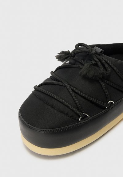 Chaussure en tissu noir avec des lacets élastiques et des pompons décoratifs, dotée d'une semelle en caoutchouc lisse de couleur beige contrastante.