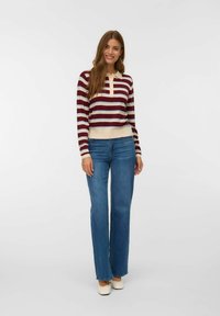 Jonge vrouw die glimlacht, in een gestreepte bordeauxrode en beige longsleeve shirt, blauwe wijde jeans en roomkleurige ballerina's, staand.