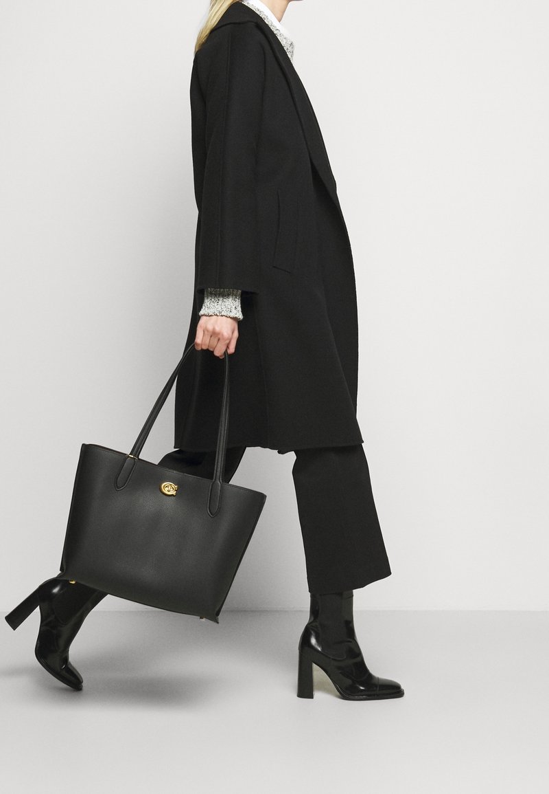 Femme en manteau et pantalon noirs marchant, portant un grand sac à main en cuir noir et des bottines noires à talons.