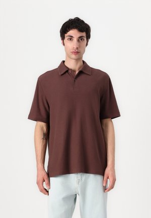 Brown short-sleeve polo shirt with a soft texture, two-button placket, and subtle logo on the chest; paired with light blue jeans.