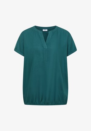 Femme aux cheveux courts et bruns portant un chemisier à manches courtes de couleur sarcelle et un jean bleu clair, vue de dos sur fond beige.