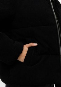 Sort kordfløjl puffjakke med en struktureret overflade, der har lynlås, sidelommer og ribbetræksmanchetter for ekstra varme.