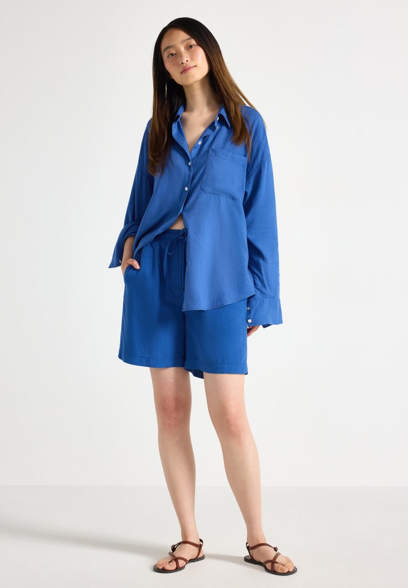Camicia blu con maniche lunghe e taschino frontale, abbinata a pantaloni corti blu coordinati. Tessuto leggero con vestibilità comoda. Sandali marroni con cinturini.