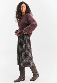 Bordowy, puszysty sweter, spódnica w kratę z bordowym i zielonym wzorem, brązowe zamszowe kozaki za kolano z detalem w postaci klamry. Elegancki, warstwowy strój.