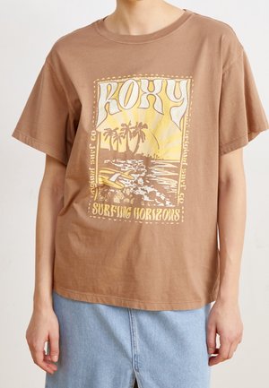 Personne portant un t-shirt marron à manches courtes avec une scène de surf jaune, des palmiers et le texte "ROXY Surfing Horizons" sur le devant.