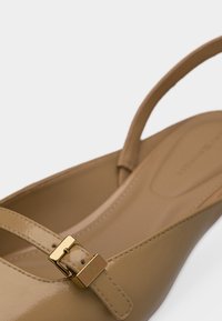 Μπεζ γυαλιστερό δερμάτινο παπούτσι με λουρί πίσω, λεπτό λουράκι και μικρή χρυσή αγκράφα σε λευκό φόντο.