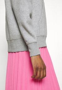 Grå sweatshirt med ribbade muddar och fåll tillsammans med en plisserad rosa kjol. Strukturerat tyg syns på båda plaggen.