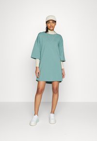 Vestido t-shirt oversized teal com mangas curtas, feito de tecido suave. Estilizado com ténis brancos e um gorro leve bege. Design simples.