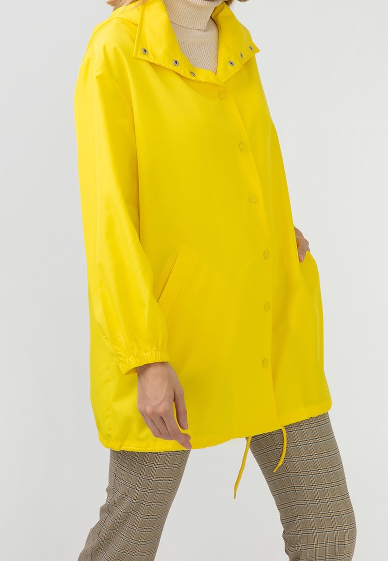 Personne portant un imperméable jaune vif sur un col roulé beige et un pantalon à carreaux, debout les mains dans les poches devant un fond uni.