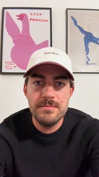 Λευκό καπάκι μπέιζμπολ με μπορντό λεπτομέρειες, που φέρει το κείμενο "Απλά κάν' το" σε καφέ χρώμα. Το μοντέλο φοράει μια μαύρη σαλοπέτα, πλαισιωμένο από πολύχρωμες τέχνες εκτυπώσεις.