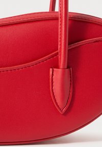 Sac à main en cuir rouge avec une forme arrondie, une texture lisse et une fine bandoulière ornée d'un accent décoratif au niveau de la fixation de la sangle.