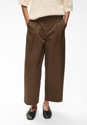 Personne portant un pantalon marron ample coupé court, un pull en tricot beige clair et des chaussures plates noires, les mains dans les poches.