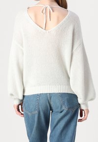 Bílý pletený svetr s hlubokým výstřihem na zádech a detailem na zavazování. Má oversized rukávy a žebrovaný lem. Spárován s modrými džíny.