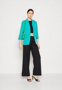 Blazer turquesa de corte slim, top negro corto, pantalones negros de pierna ancha y tacones abiertos negros. Incluye un bolso verde texturizado.