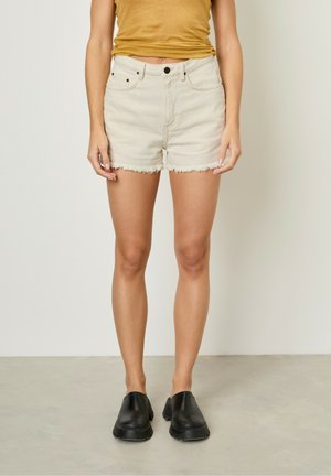 Shorts en denim beige avec un ourlet effiloché, design à cinq poches et fermeture à bouton noir, associés à un débardeur moulant de couleur moutarde et des chaussures noires.