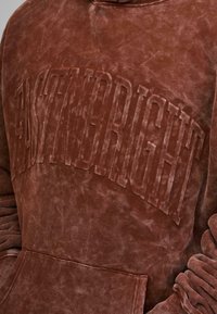 Felpa marrone realizzata in materiale morbido, con un logo in rilievo e testurizzato sulla parte anteriore. Il tessuto ha un aspetto vintage e sbiadito.