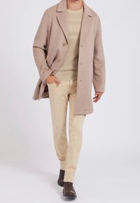 Mand der går i beige frakke, stripet striktrøje, lysebeige bukser og mørkebrune læderstøvler mod en ensfarvet lys baggrund.