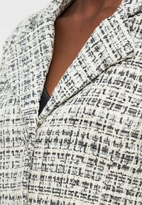 Μαύρο και λευκό υφασμάτινο σακάκι από tweed με φαρδιές πέτες πάνω από μαύρη μπλούζα, αποκαλύπτοντας μερικώς την περιοχή του στήθους.