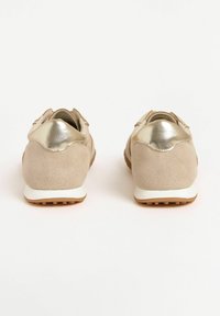 Sneaker beige con un accento dorato lucido sul tallone, viste da dietro, appoggiate su uno sfondo chiaro.
