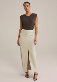 Brun ärmlös topp, högmidjad ljusbeige kjol med sidofålla, och svarta sandaler med remmar. Slät tyg med en subtil struktur.