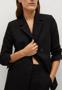 Žena v černém dvouřadovém blejzru a odpovídajících kalhotách, držící klopu blejzru jednou rukou a druhou ruku má v kapse.