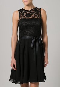 Robe noire avec un corsage en dentelle, conception sans manches et jupe fluide. Elle présente un ruban en satin noir à la taille. La dentelle a un motif floral.