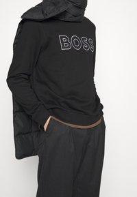 Černá mikina s logem "BOSS", k tomu tmavá péřová vesta přehozená přes rameno, doplněná šedými kalhotami s hnědým páskem.