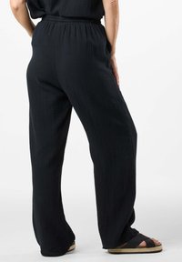 Pantalon noir à jambes larges en tissu texturé, avec une taille élastique et des poches latérales, associé à des sandales noires à talons plats.