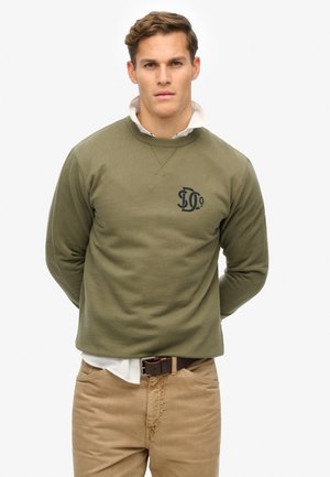 Olijfgroene crewneck sweatshirt met ribbelstructuur, met een donker logo op de borst, gecombineerd met beige broek en een witte overhemd eronder.