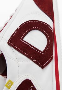 Adidași albi cu accente de suede roșu și cusături. Logo-ul tăiat notabil pe lateral și eticheta galbenă aproape de călcâi. Talpă din cauciuc.