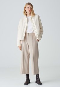 Veste matelassée beige avec des boutons pression, associée à un pantalon large beige taille haute et des bottes marron à talons montantes. Chemise blanche en dessous.