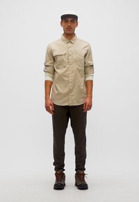 Άνδρας που φοράει μπεζ μακρυμάνικο πουκάμισο, σκούρο παντελόνι, ορειβατικά μποτάκια και λευκό καπέλο, στέκεται μπροστά σε λευκό, μονόχρωμο φόντο.