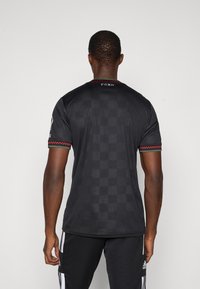 Camiseta deportiva negra con un patrón de tablero de ajedrez texturizado, acentuada con ribetes rojos y verdes a lo largo del collar y las mangas.