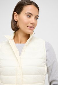 Elfenbeinfarbene gesteppte Weste mit hohem Kragen und Reißverschluss vorne. Wird über einem hellgrauen Pullover getragen. Dezente Markenkennzeichnung auf der Brust.