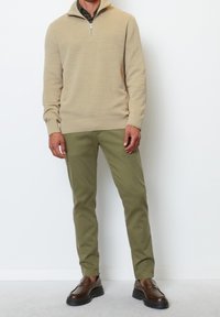 Homme portant un pull beige à fermeture éclair quart, pantalon vert olive et mocassins marron foncé, debout contre un mur blanc uni.