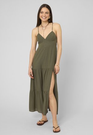Femme souriante dans une robe maxi verte sans manches avec une fente, portant des colliers superposés, un bracelet, une chaîne de cheville et des sandales plates marron.