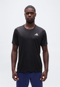 Άνδρας που φοράει μαύρο αθλητικό t-shirt της Adidas και μπλε σορτς, στέκεται μπροστά σε απλό ανοιχτό γκρι φόντο, κοιτώντας μπροστά με ουδέτερη έκφραση.