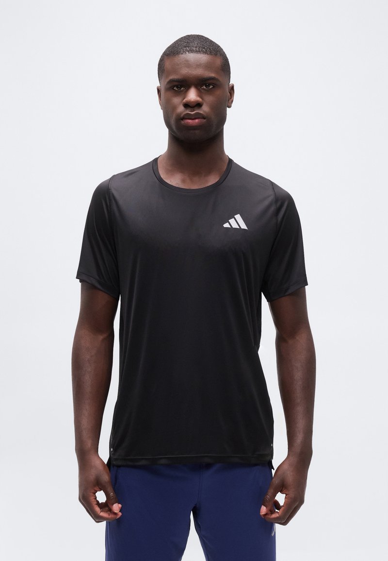 Άνδρας που φοράει μαύρο αθλητικό t-shirt της Adidas και μπλε σορτς, στέκεται μπροστά σε απλό ανοιχτό γκρι φόντο, κοιτώντας μπροστά με ουδέτερη έκφραση.