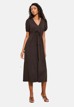 Femeie purtând o rochie midi neagră cu mâneci scurte bufante, decolteu în V și cordon în talie, asortată cu sandale maro cu toc și o brățară.