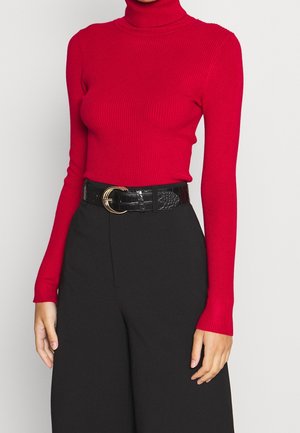 Vrouw draagt een aansluitende rode geribbelde coltrui, ingestopt in een zwarte broek met hoge taille, met een zwarte riem met een gouden dubbelringgesp.